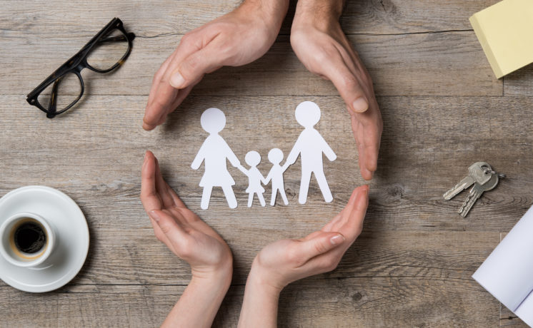 Close up of female and male hands protecting a paper chain family. Top view of two hands form a circle around white paper chain family on wooden table. Family care, insurance and helping hand concept.